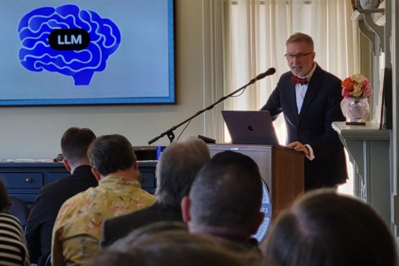 Man in suit and bowtie stands behind a lecturn with a screen behind him featuring an illustration of a brain and the letters LLM.