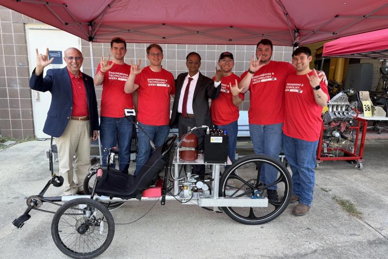 Seven people stand in front of a hydraulic vehicle.