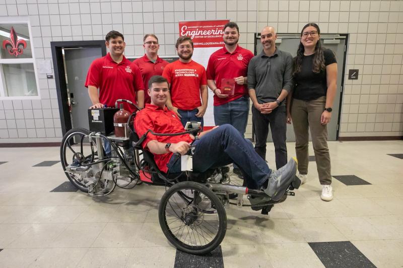 Six people stand for a group photo with one person sitting in a tricycle holding a trophy.