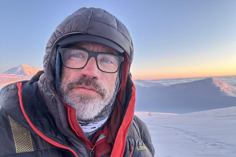 Man in climbing gear stands on top of a mountain covered in snow
