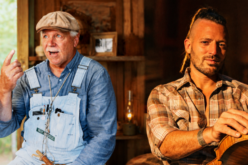 Two men sitting in a living room. One hands a fiddle, the other wears a duck call around his neck.