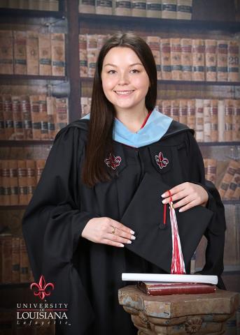 Jenna Pryor in UL Lafayette black graduation gown in front of photo backdrop of shelved books.