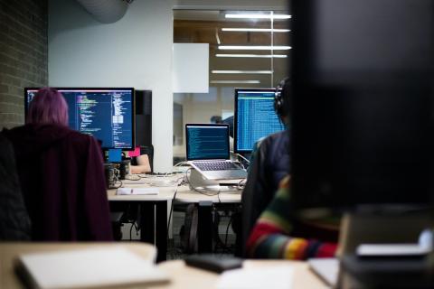 Several individuals sit at computers with code on the screens. 