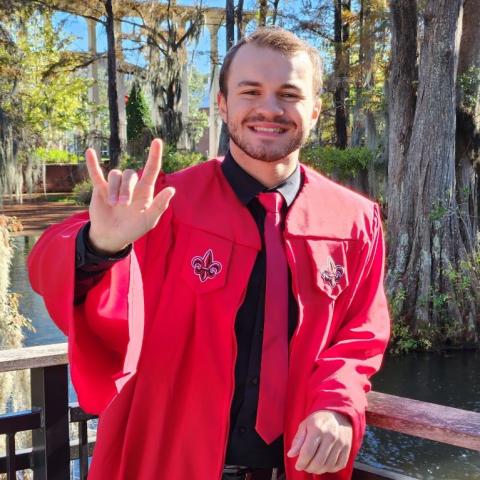 Drake Bourgeois pictured outdoors in a red, UL Lafayette graduate gown holding up his right hand in the UL symbol 