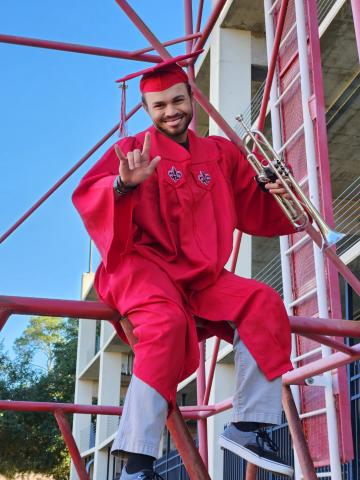 Drake Bourgeois pictured outdoors in a red, UL Lafayette graduate gown holding up his right hand in the UL symbol and holding a saxophone in his left hand.  
